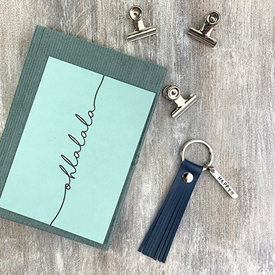 Flatlay of a mint colored notebook and postcard with text 'olllalala', 3 metal clips and a dark blue leather keychain.