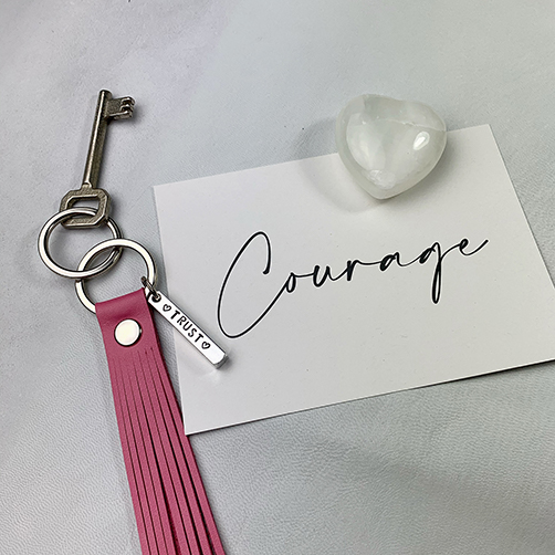 Hot pink leather tassel keychain with personalized metal bar with the word TRUST and a white postcard with the word Courage written in black.